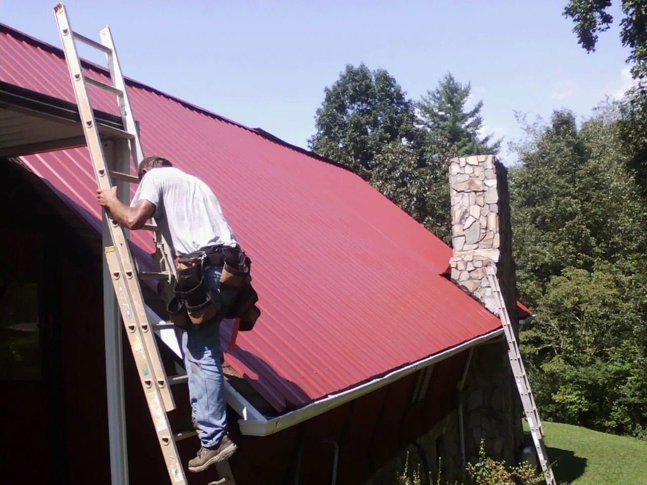 Roofer on a ladder beside a red metal roof and stone chimney — Soffit & Fascia Repair by Roofing Brothers in Lawn
